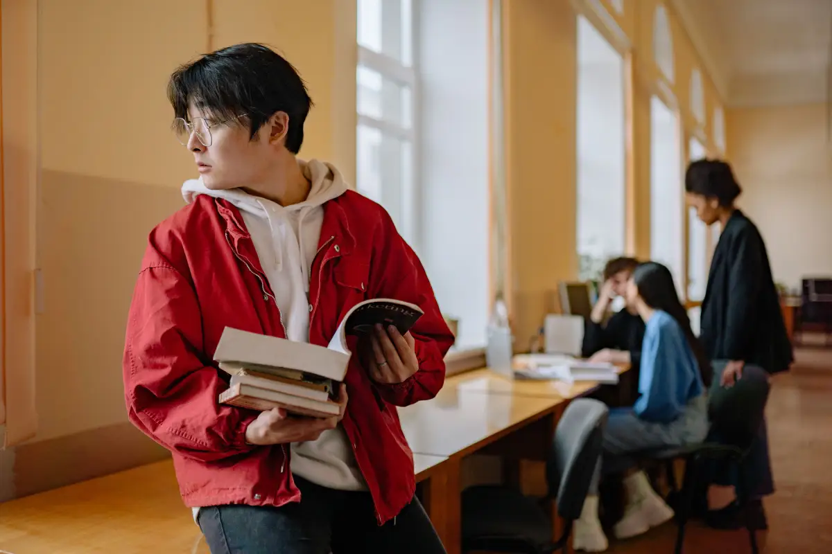 college student in red jacket holding book looking out window